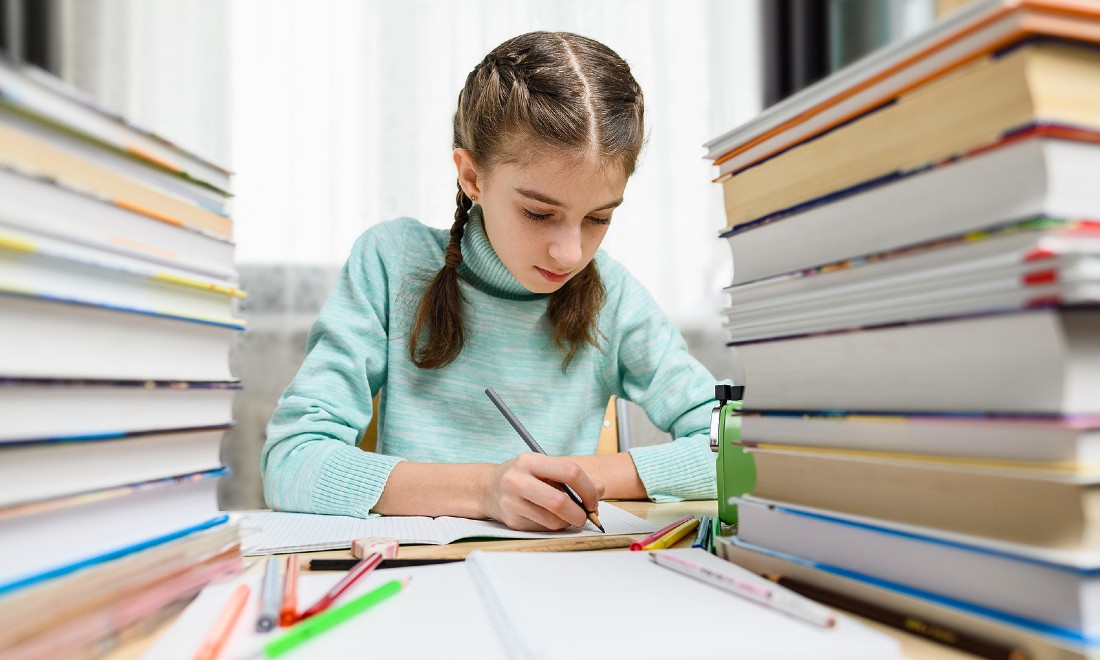 girl sitting next to book piles, doing homework