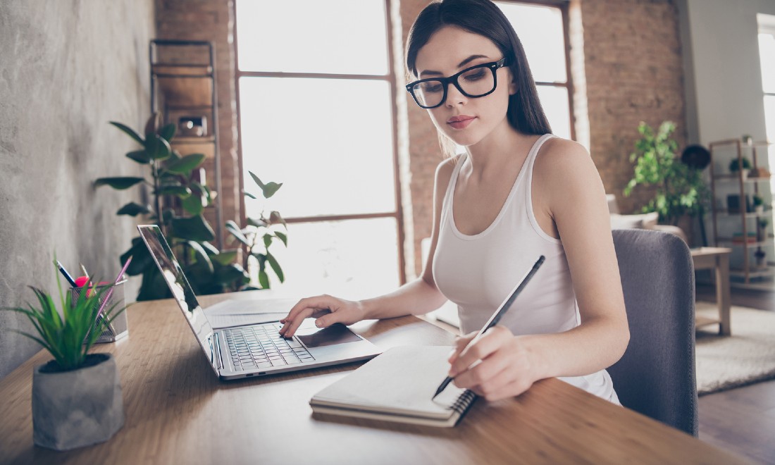 woman working from home on laptop, writing notes in notebook