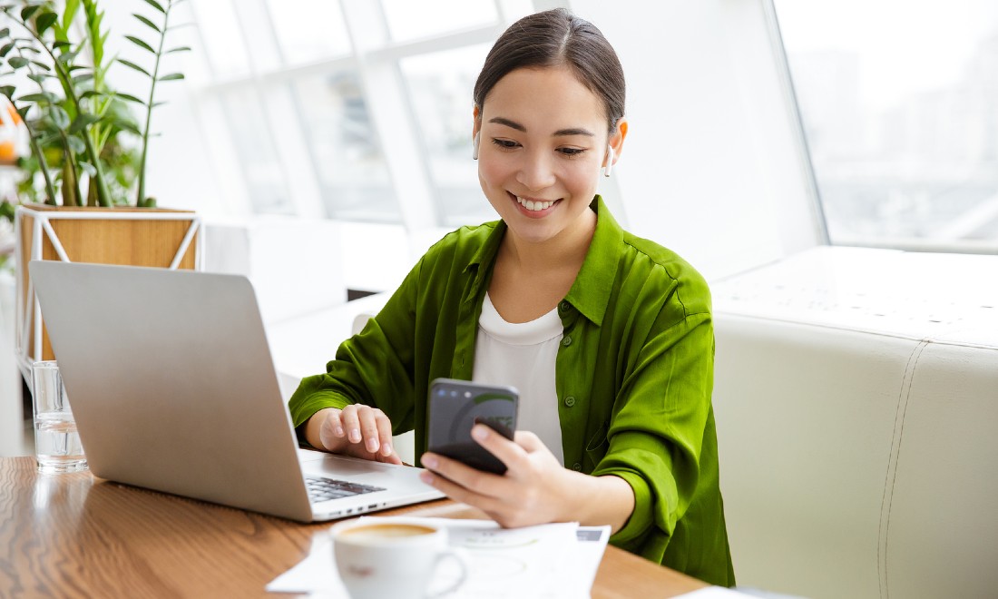 girl looking at phone while working on laptop