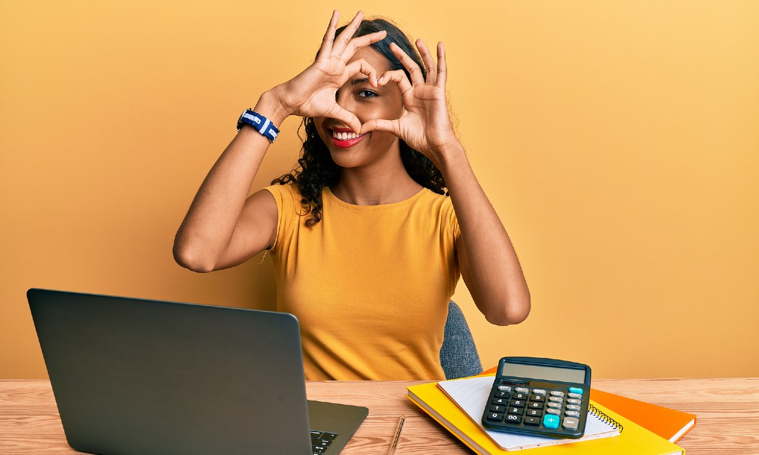 woman making heart sign with hands sitting behind desk with laptop and calculator