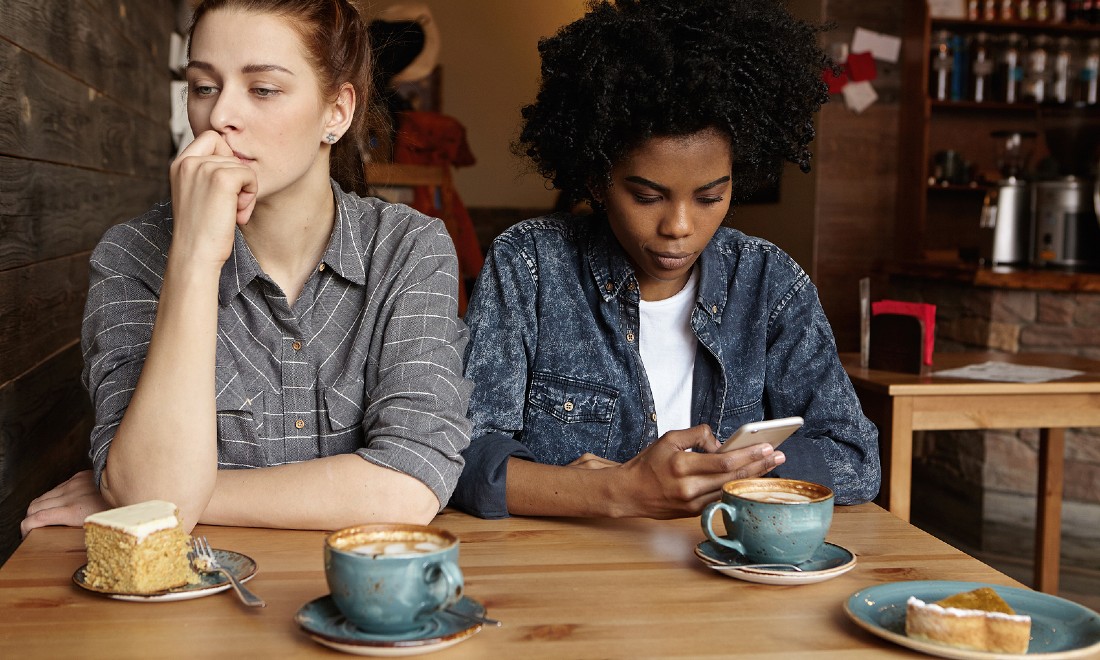 two women at table during lunch, one looks sad, the other is on phone